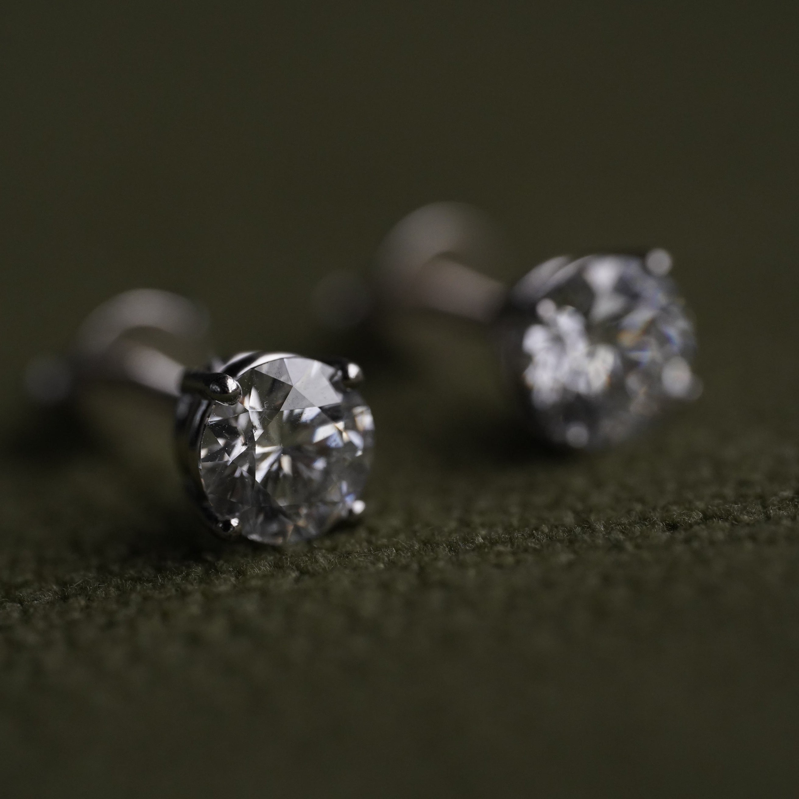 A pair of round-cut stud earrings with silver-toned settings on a dark olive green textured surface.
